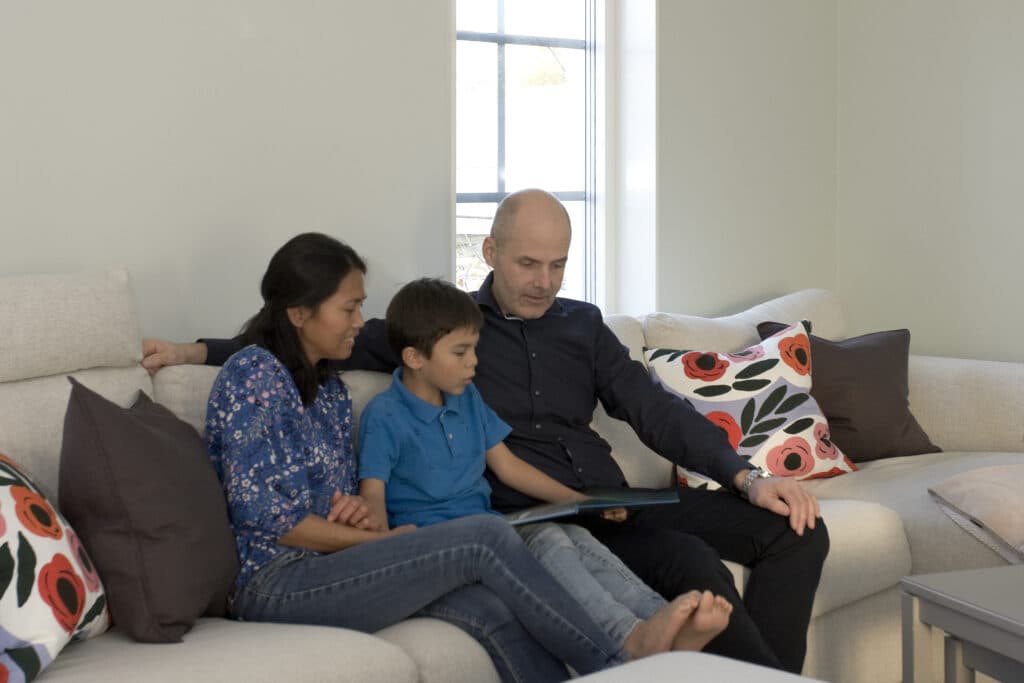 En familj sitter på en vit soffa. Pojken löser en bok och föräldrarna sitter bredvid.  